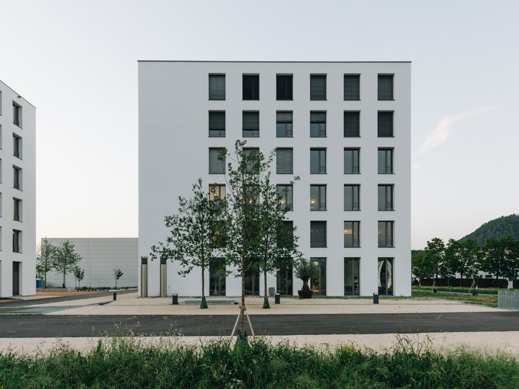 Außenansicht eines mehrstöckigen Bürogebäudes mit betonierter Außenfläche; mehrere Bäume und grüne Wiese sind im Vordergrund.